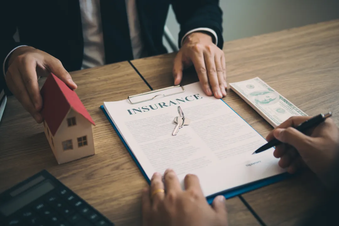 127907985_m A person signs an insurance document with a pen, alongside a small house model and a dollar bill on a wooden table.