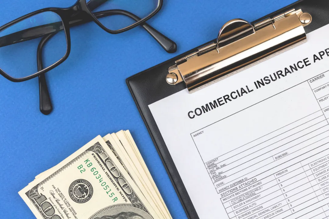 171687773_m A clipboard with a commercial insurance application, next to a stack of cash and black glasses on a blue background.